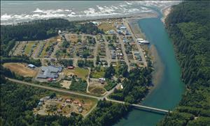 Aerial view of a small town with a river on one side and an ocean beach on the other. A bridge crosses the river towards forested hillsides. 